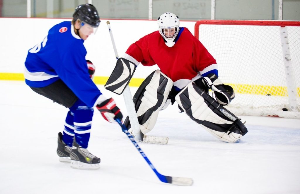 A playing in blue aims to shoot the puck at the goalie in red playing ice hockey